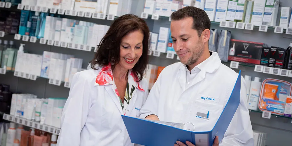 Mag. pharm. Alexander Koller und MMag. pharm. Doris Koller besprechen sich vor einem Regal der St. Anna-Apotheke